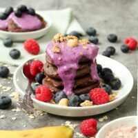 Hier kann man protein Pancakes mit Banane auf einem weißen Teller mit verschiedenen Beeren als Dekoration darauf
