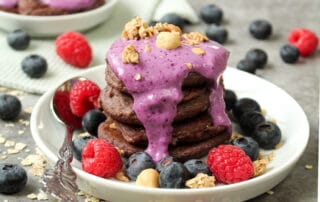 Hier kann man protein Pancakes mit Banane auf einem weißen Teller mit verschiedenen Beeren als Dekoration darauf